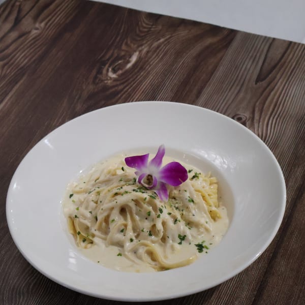 Creamy pasta dish garnished with chopped herbs and a purple orchid on a white plate.