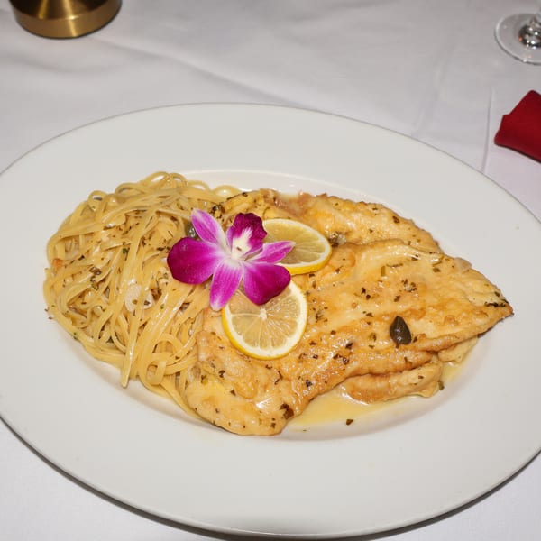 Chicken piccata with linguine, lemon slices, capers, and an orchid garnish on a white plate.